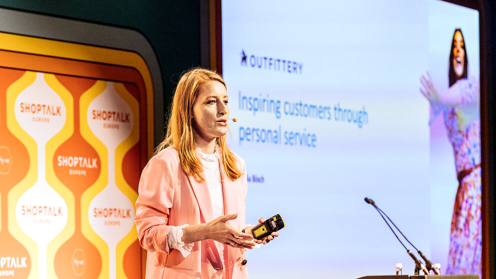 Frau in rosa Blazer hält Präsentationsfernbedienung vor Leinwand mit Text 'Inspiring customers through personal service' und Logo 'OUTFITTERY'.