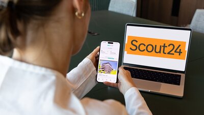 Zu sehen ist ein Laptop mit dem Logo des Unternehmens Scout24. Davor sitzt eine Frau mit hochgesteckten braunen Haaren, der man über die Schulter auf den Laptop schaut.