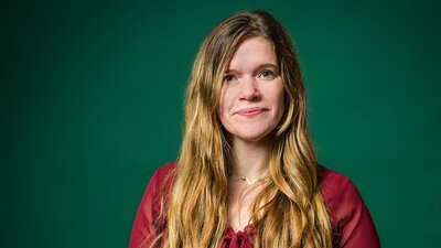 Zu sehen ist eine Frau mit rotem Oberteil und blonden langen Haaren. Sie steht vor einer grünen Wand und schaut neutral in die Kamera.