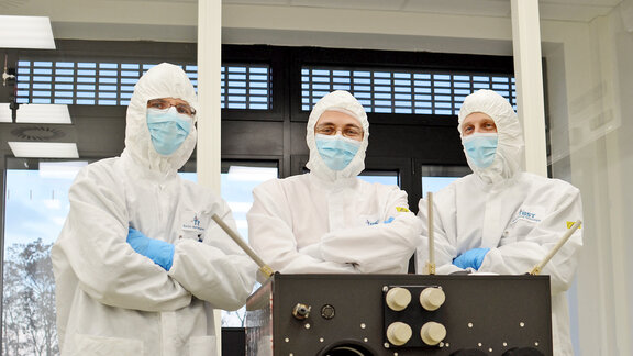 Drei Personen in weißen Schutzanzügen und blauen Handschuhen stehen hinter einem technischen Gerät mit mehreren Knöpfen und Hebeln in einem hellen Raum.
