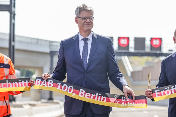 Mann in Anzug hält ein rotes Band mit der Aufschrift 'Eröffnung BAB 100, Berlin'. Zwei Personen stehen daneben, eine in orangefarbener Arbeitskleidung.