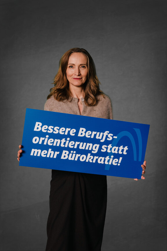 Eine Person steht vor einem neutralen grauen Hintergrund und hält ein großes blaues Schild mit der weißen Aufschrift: "Bessere Berufsorientierung statt mehr Bürokratie!"