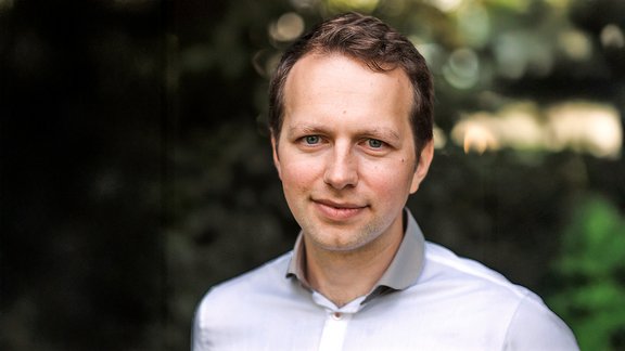 Mann mit braunen Haaren trägt ein weißes Hemd, unscharfer grüner Hintergrund mit Lichtflecken.