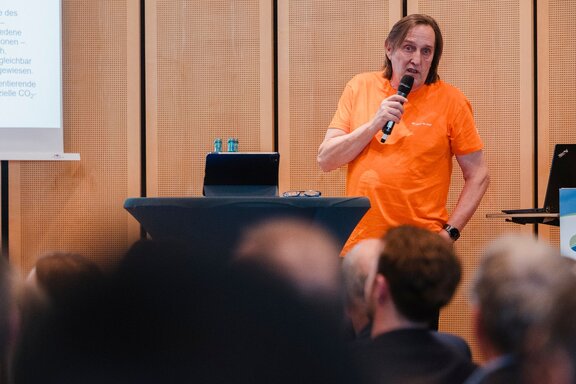 Zu sehen ist ein Mann mit mittellangen Haaren und einem orangenem Shirt, welcher auf einer Bühne vor Publikum in ein Mikrofon spricht.