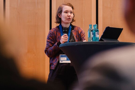 Zu sehen ist ein junge Frau, die auf einer Bühne vor Publikum in ein Mikrofon spricht.