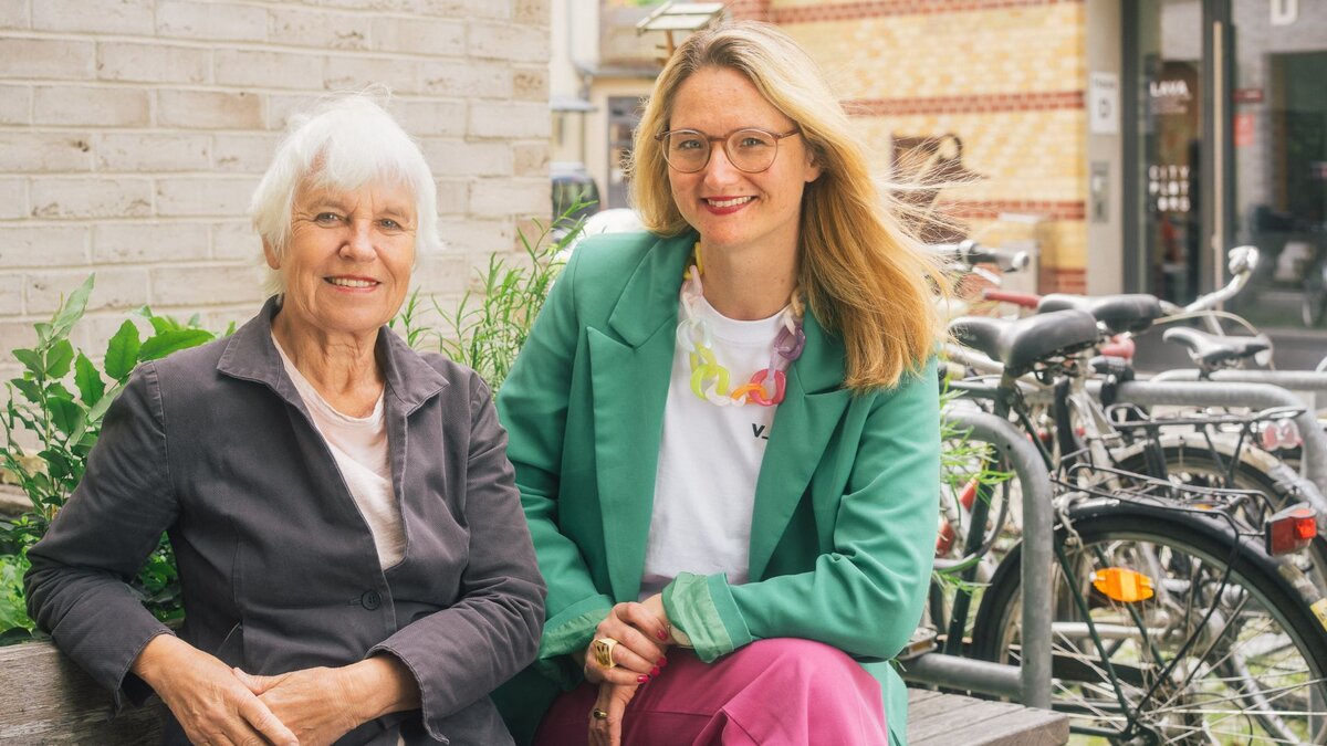Zu sehen ist eine ältere Frau mit kurzen weißen Haaren und eine mittelalte Frau mit langen blonden Haaren. Die Damen sitzen nebeneinander auf einer Bank, im Hintergrund Fahrräder und sie schauen freundlich in die Kamera. Es handelt sich um velo-konzept-Gründerin Ulrike Saade und ihre Nachfolgerin Isabell Eberlein.