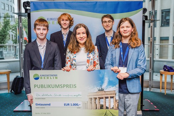 Fünf junge Menschen stehen zusammen und halten einen großen Scheck mit der Aufschrift 'Publikumspreis' und 'EUR 1.000' vor einem Banner von Klimaschutzpartner Berlin.