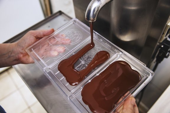 Zwei Hände halten eine transparente Form, in die flüssige Schokolade aus einem Metallrohr gegossen wird.