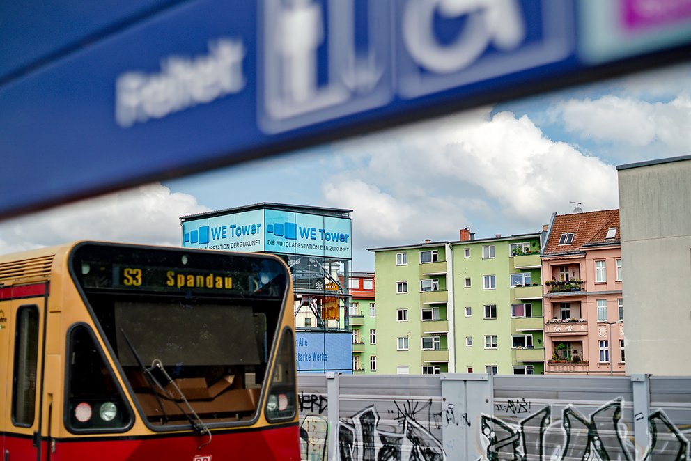 Ein roter Zug mit gelben Akzenten fährt an einem Bahnhof vorbei. Im Hintergrund sind mehrstöckige, farbige Wohngebäude zu sehen. Ein Schild mit 'WE Tower' ist sichtbar.