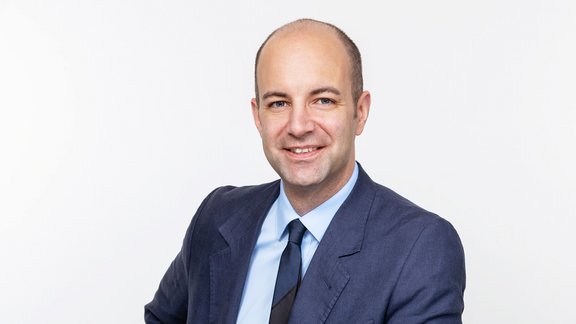 Mann mit Glatze trägt dunkelblauen Anzug, hellblaues Hemd und dunkelblaue Krawatte vor weißem Hintergrund.