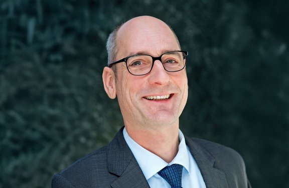 Oberkörper eines Mannes mit grauen Haaren, Brille, weißem Hemd, blauem Krawattenmuster und dunkelgrauem Sakko vor unscharfem Hintergrund.