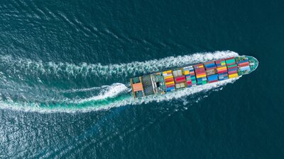 Zu sehen ist ein Containerschiff, welches von oben im Meer fotografiert wurde.