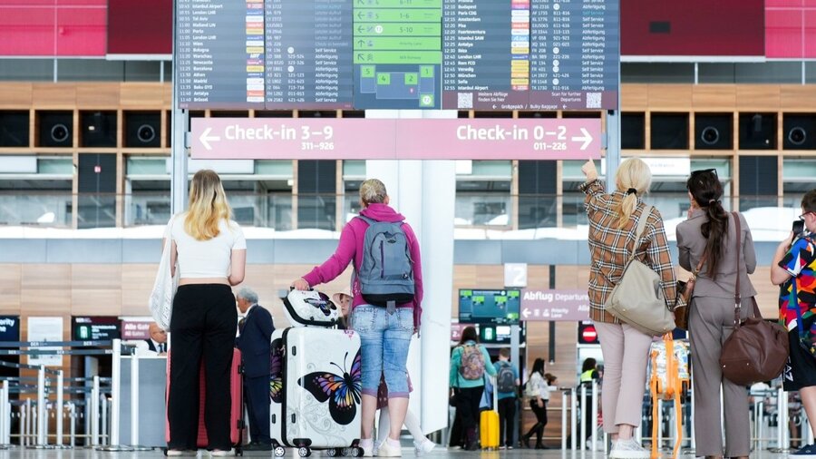 Zu sehen sind vier Personen von hinten. Sie stehen in einem Flughafen neben ihren Koffern und schauen auf eine Anzeigetafel.