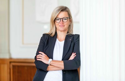 Frau mit blonden Haaren, verschränkten Armen, trägt schwarzen Blazer und weiße Bluse, steht vor weißer Wand mit Säule