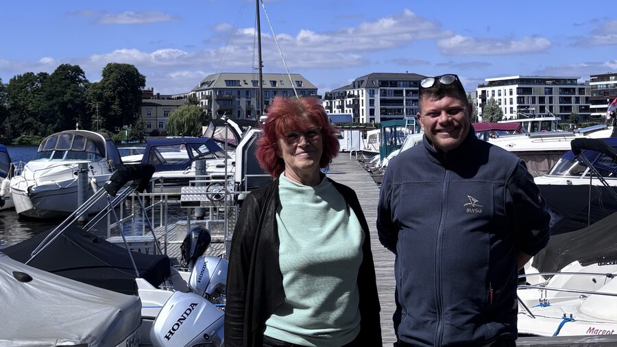 Zu sehen sind eine Frau und ein Mann am Steg eines Gewässers, hinter ihnen sind verschiedene Boote zu erkennen.