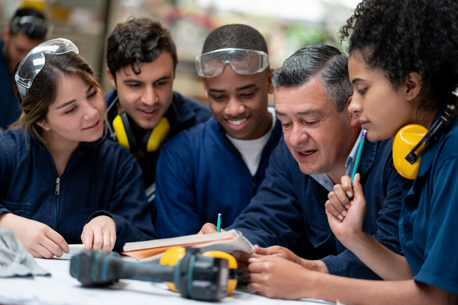 Zu sehen sind mehrere Arbeitende, in dunkelblauen Overalls, Zwei von ihnen tragen eine Schutzbrille, zwei weitere haben Lärmschutzkopfhörer um den Hals hängen. Eine Person scheint der Ausbilder zu sein und erklärt den restlichen Arbeitenden, die um einen Tisch stehen etwas. Auf dem Tisch liegt ein Akkuschrauber.