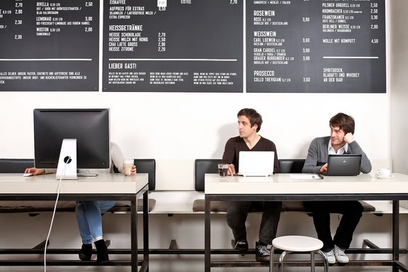 Drei Personen sitzen an Tischen mit Laptops und Monitoren, darüber hängt eine Speisekarte mit Getränkepreisen in deutscher Sprache