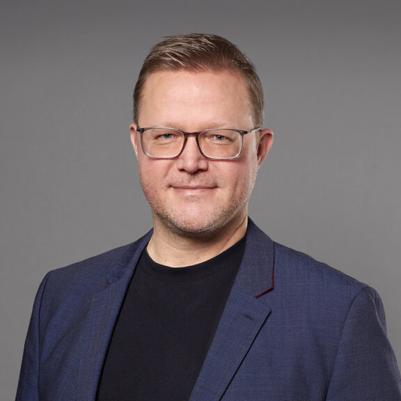 Auf dem Bild ist ein Mann mit Brille und blauem Jackett zu sehen, der in die Kamera guckt.