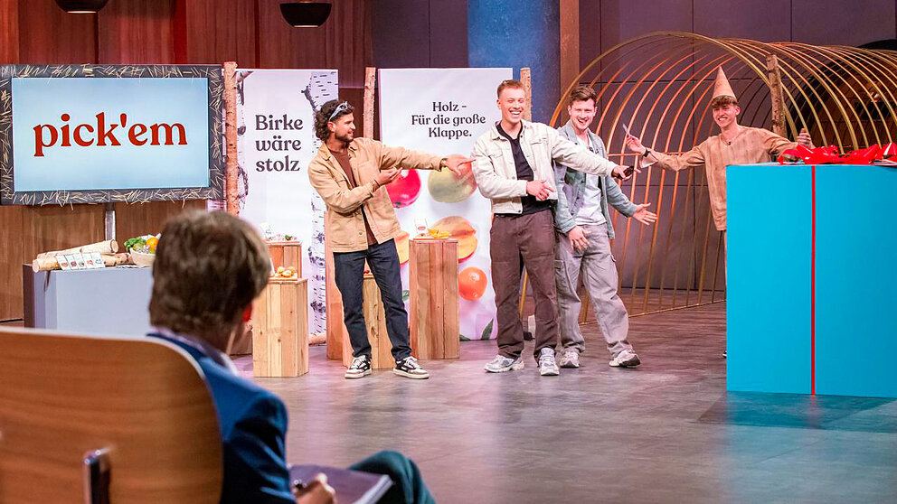 Vier Männer stehen auf einer Bühne, zwei halten Schilder mit den Texten 'Birke wäre stolz' und 'Holz – Für die große Klappe'. Ein Mann sitzt im Vordergrund auf einem Stuhl und notiert etwas.