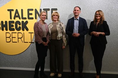 Zu sehen sind drei Frauen und ein Mann, die vor einer glitzernden silbernen Wand stehen, auf der ein gelber Kreis und der Schriftzug Talente Check Berlin stehen.