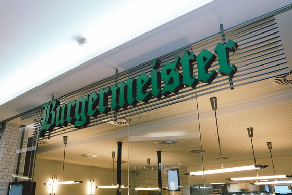 Grünes Leuchtschild mit dem Schriftzug 'Burgermeister' an einer Ladenfassade in moderner Einkaufsumgebung