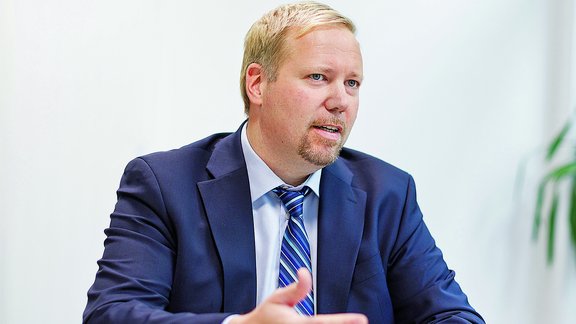 Ein Mann in einem Anzug mit Krawatte sitzt an einem weißen Tisch und gestikuliert mit den Händen. 
