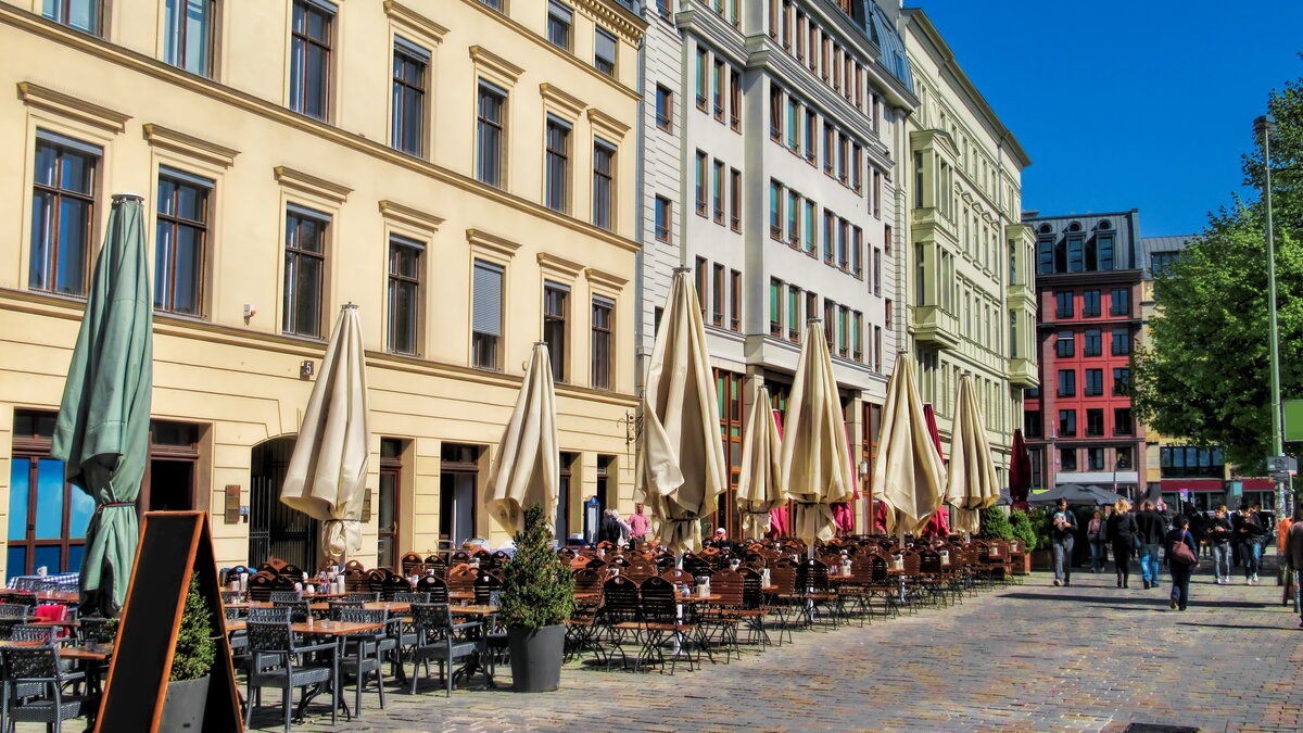 Outdoor‑Sitzbereich mehrerer Cafés und Restaurants in einer sonnigen europäischen Innenstadtstraße; lange Reihen von Tischen und Stühlen mit geschlossenen Sonnenschirmen, flankiert von historischen mehrstöckigen Gebäuden und belebt von zahlreichen Passanten im Hintergrund.