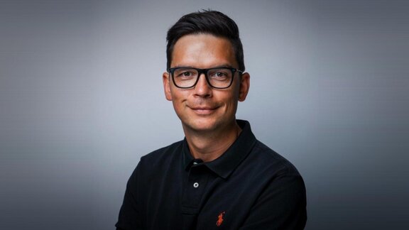 Ein lächelnder jüngerer Mann mit Brille und dunklem Oberhemd vor einem grauen Hintergrund.