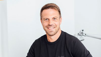 Mann mit kurzen blonden Haaren trägt ein schwarzes Calvin Klein T-Shirt, sitzt vor weißer Wand mit Handtuchhalter im Hintergrund.