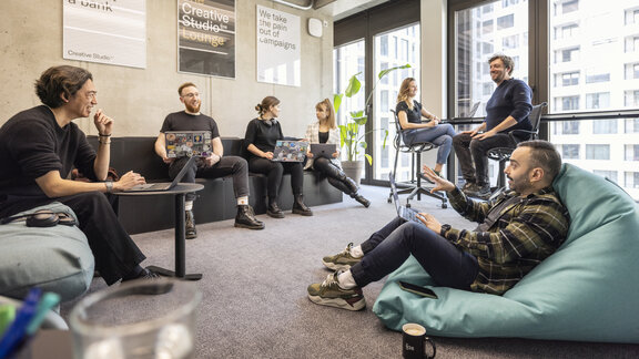 Arbeitnehmer sitzen in einem offenen, hellen Büro. Sie unterhalten sich und arbeiten an Laptops. 