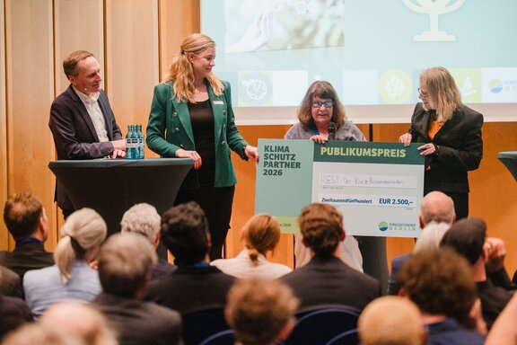 Zu sehen ist eine Bühne, darauf vier Personen. Eine Dame erhält einen überdimensionalen Check.