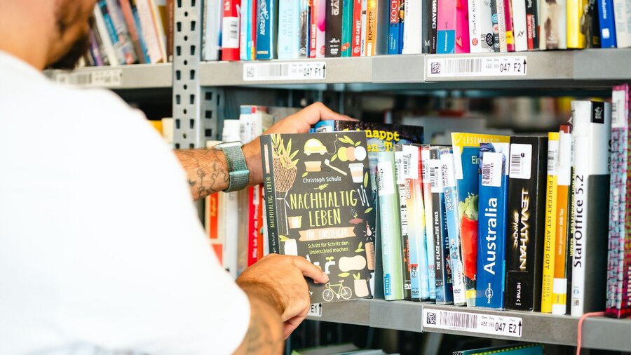 Zu sehen ist ein volles Bücherregal, aus dem eine Person gerade ein Buch herauszieht.