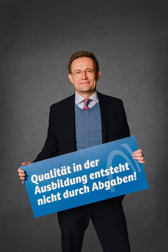 Eine Person steht vor einem neutralen grauen Hintergrund und hält ein großes blaues Schild mit der weißen Aufschrift: "Qualität in der Ausbildung entsteht nicht durch Abgaben!"