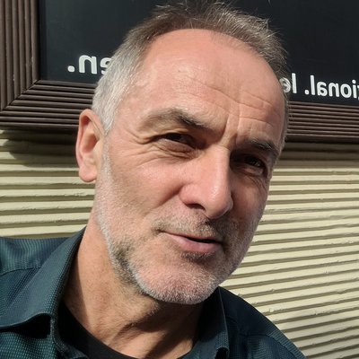 Älterer Mann mit grauen Haaren und Hemd vor einer Wand mit horizontalen Lamellen und teilweise sichtbarem Text.