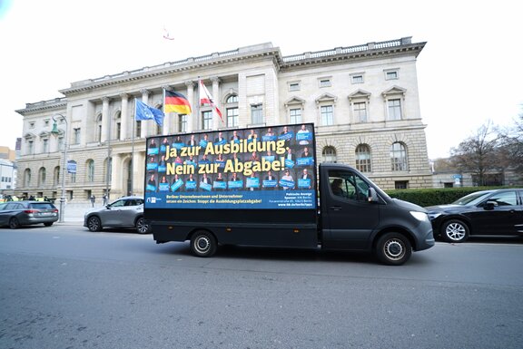 Zu sehen ist ein Transporter mit einer Werbetafel, welcher vor dem Berliner Abgeordnetenhaus steht oder vorbeifährt.