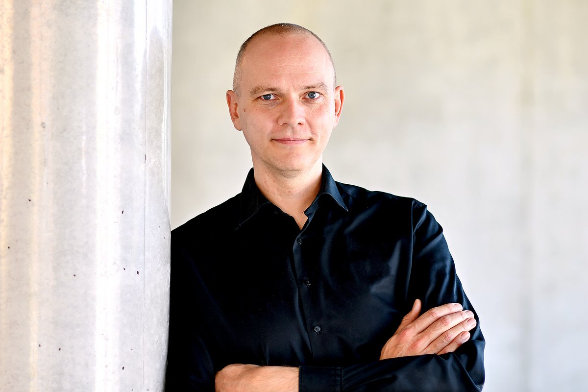 Ein Mann lehnt mit verschränkten Armen an einer hellen Betonwand und trägt ein schwarzes Hemd.