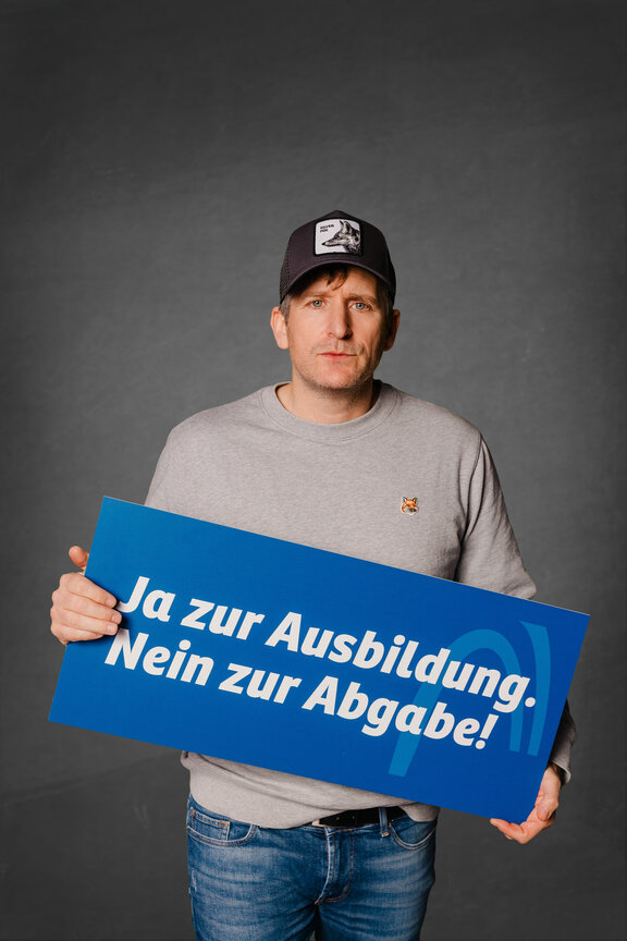 Eine Person steht vor einem neutralen grauen Hintergrund und hält ein großes blaues Schild mit der weißen Aufschrift: "Ja zur Ausbildung. Nein zur Abgabe!"