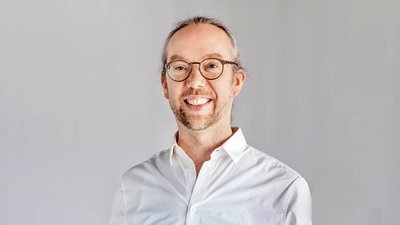 Person mit grauem Haar, zurückgebunden, trägt ein weißes Hemd mit offenem Kragen vor neutralem Hintergrund