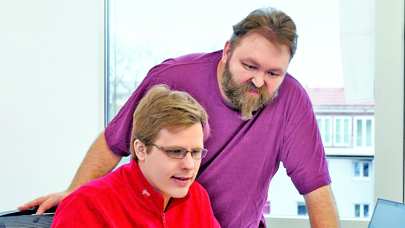 Zwei Männer blicken auf einen Computerbildschirm, einer sitzt am Schreibtisch