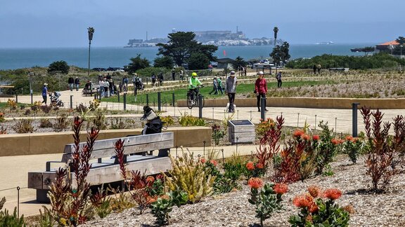 Ein Park mit Holzbänken, großen Grünflächen und vielen Pflanzen. Auf den Gehwegen laufen Menschen. 