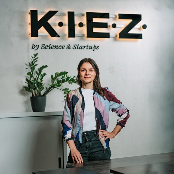 Frau steht mit einer Hand in der Hüfte vor Wand mit beleuchtetem Schriftzug 'K.I.E.Z. by Science & Startups', trägt bunte Jacke und weiße T-Shirt