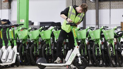 Zu sehen ist ein junger Mann, welcher eine hellgrüne Warnweste trägt und einen weiß-grünen E-Roller technisch überprüft.