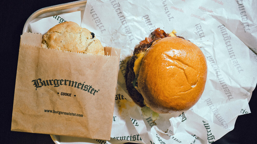 Burgermeister Cheeseburger mit geschmolzenem Käse und ein großer Cookie in einer Papiertüte mit Burgermeister-Logo
