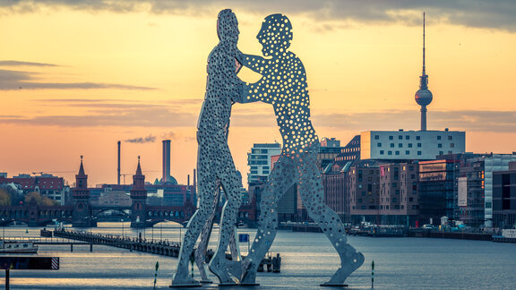 Zu sehen ist eine große metallene Statue von zwei tanzenden Personen. Die Statue befindet sich im Wasser. Es ist der Molecule-Man in der Spree in Berlin. Im Hintergrund sieht man die Stadt mit dem Fernsehturm als Wahrzeichen.