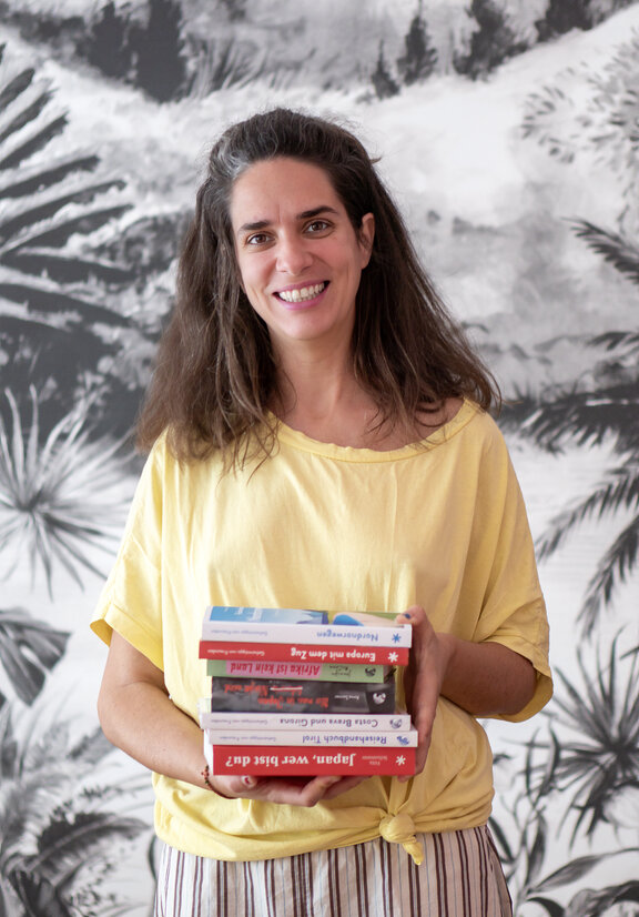 Eine Frau mit einem gelben T-Shirt steht vor einer schwarz/weißen Tapete, die ein tropisches Muster hat. In der Hand hält Sie einen Stapel Bücher