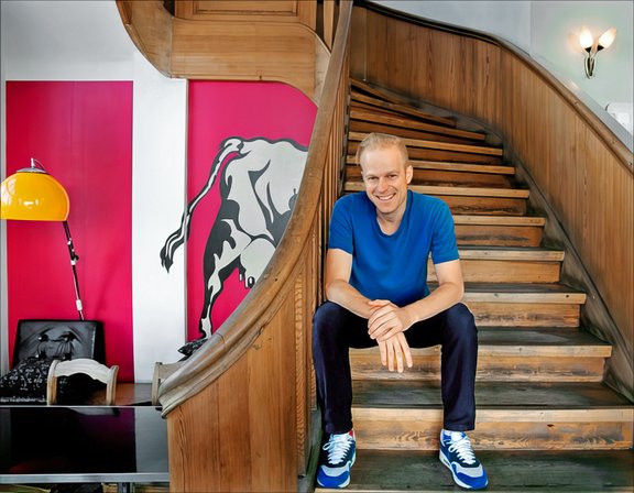 Mann in blauem T-Shirt und Jeans sitzt auf einer Holztreppe in einem Raum mit rotem Wandbild eines Stiers und gelber Stehlampe