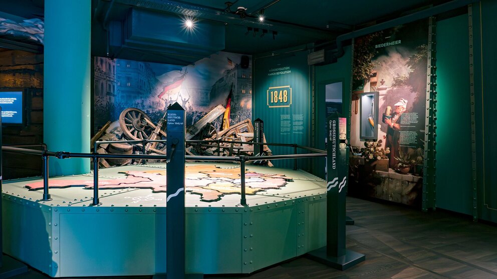 Zu sehen ist der Innenraum eines Museums. Im Zentrum eine Erhöhung, auf der eine Landkarte zu sehen ist. Im Hintergrund ein Schild mit der Aufschrift 1848. Es handelt sich um das Deutschlandmuseum.