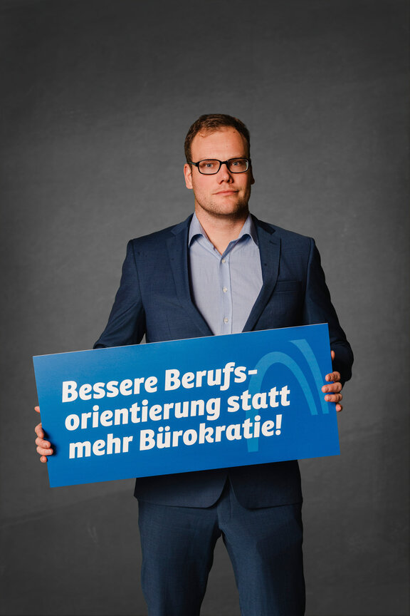 Eine Person steht vor einem neutralen grauen Hintergrund und hält ein großes blaues Schild mit der weißen Aufschrift: "Die Strafabgabe schwächt den Wirtschaftsstandort!"