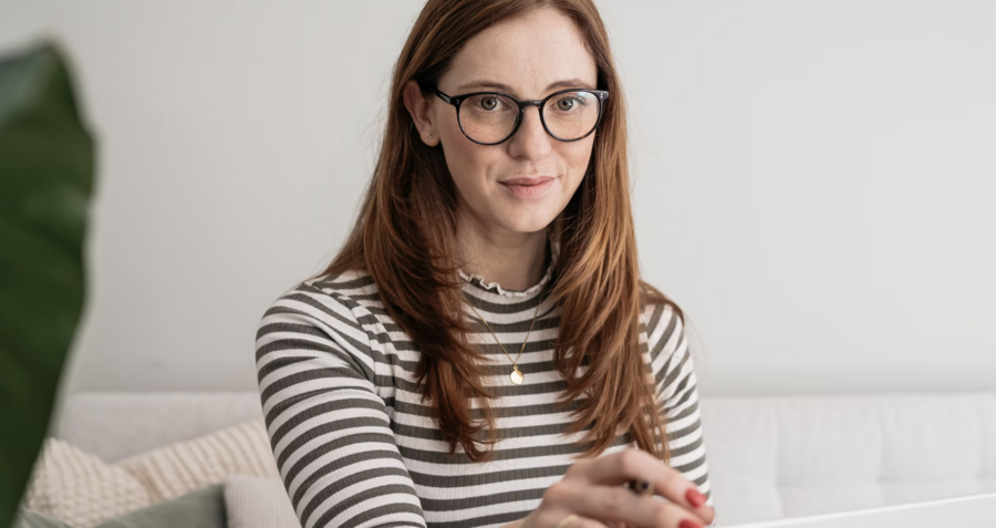 Zu sehen ist eine junge Frau mit langen braunen Haaren, einer Brille und einem schwarz-weiß-gestreiftem Shirt.