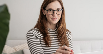 Zu sehen ist eine junge Frau mit langen braunen Haaren, einer Brille und einem schwarz-weiß-gestreiftem Shirt.